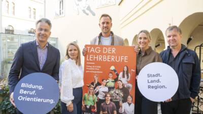 Fünf Personen präsentieren ein orangefarbenes Plakat zum Tag der Lehrberufe und zwei runde Schilder.