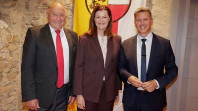 Drei Personen in formeller Kleidung stehen vor einer Fahne mit dem Wappen Kärntens.