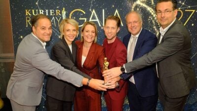 Sechs Personen in eleganter Abendkleidung halten gemeinsam eine goldene Trophäe vor einem sternenbedeckten Hintergrund.