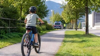 Junge mit Helm auf blauem Fahrrad faehrt einen Weg entlang mit Polizeifahrzeug im Hintergrund