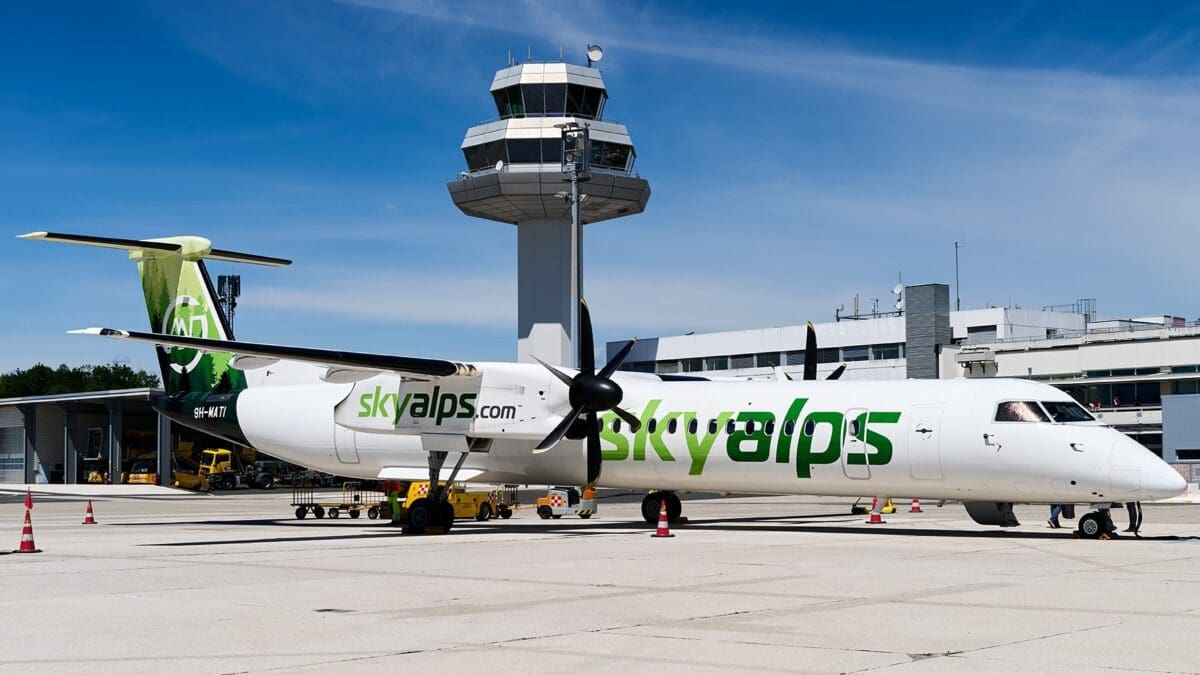 Ein weißes SkyAlps-Propellerflugzeug steht auf dem Rollfeld des Klagenfurt Airports vor dem Kontrollturm.
