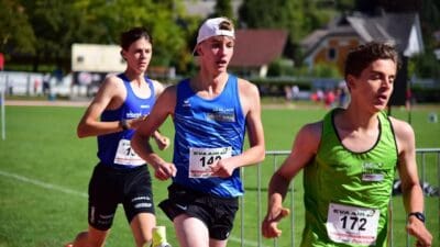 Drei junge Läufer in farbigen Trikots laufen auf einer grünen Sportwiese bei einem Wettkampf.