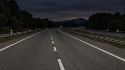 Leere Autobahn bei Nacht mit weißen Markierungen und Leitplanken zwischen bewaldeten Hängen.