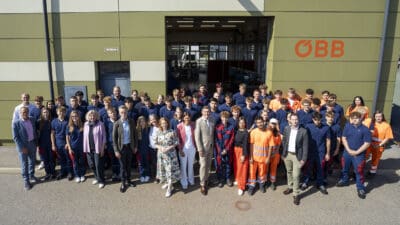 Große Gruppe junger Menschen in blauer und orangener Arbeitskleidung sowie Erwachsene in Geschäftskleidung vor einem ÖBB-Gebäude.