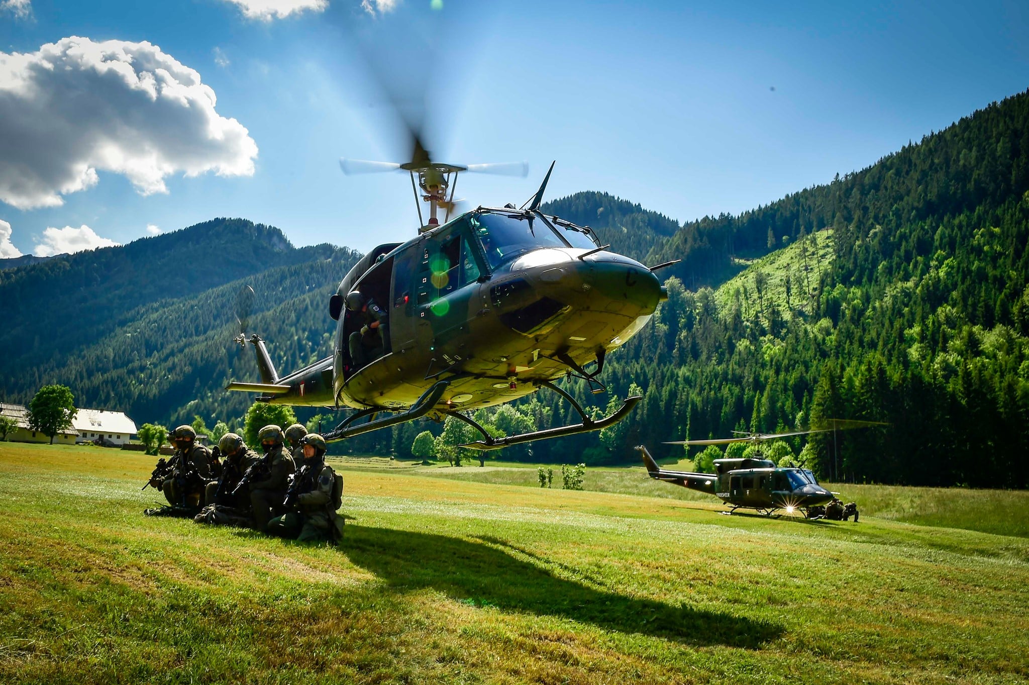 Soldaten in militärischer Ausrüstung sitzen vor einem schwebenden Hubschrauber auf einer Wiese mit Bergen im Hintergrund.
