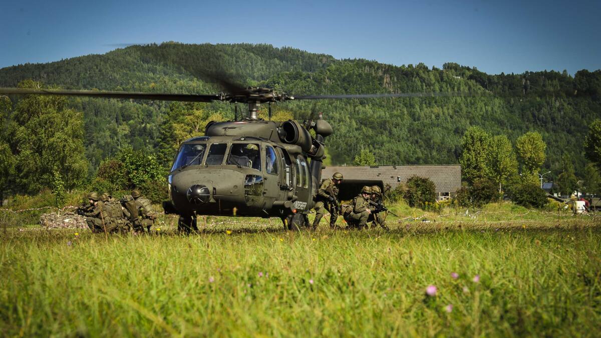 Ein Militärhubschrauber mit laufenden Rotoren steht auf einer Wiese, während Soldaten in Tarnkleidung daneben in Position gehen.