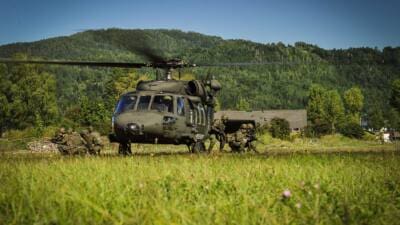 Ein Militärhubschrauber mit laufenden Rotoren steht auf einer Wiese, während Soldaten in Tarnkleidung daneben in Position gehen.