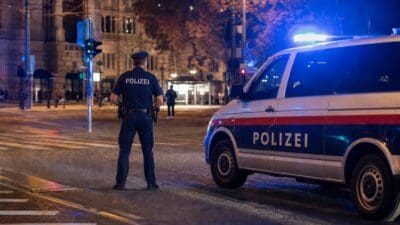 Polizist mit POLIZEI-Rückenaufschrift steht neben Streifenwagen mit Blaulicht auf nasser Straße bei Nacht