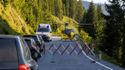 Gelber ÖAMTC-Hubschrauber steht hinter einer Absperrung auf einer Alpenstraße mit Rettungswagen und Autos.