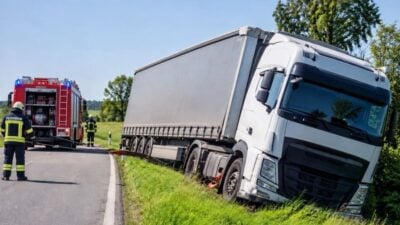 Weißer Lkw mit Anhänger steht schräg am Straßenrand in einer Böschung, im Hintergrund rotes Feuerwehrauto und Einsatzkräfte