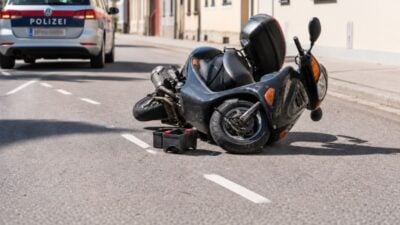 Ein umgestürzter schwarzer Motorroller liegt auf der Straße neben einem Polizeiauto mit Blaulicht.