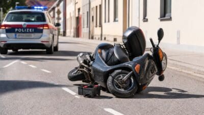 Umgestürzter schwarzer Roller liegt auf der Straße neben einem Polizeiauto mit Blaulicht.