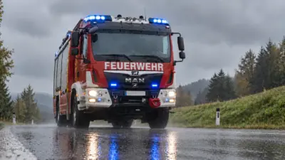 Rotes Feuerwehrfahrzeug mit Blaulicht auf regennasser Straße vor Waldhintergrund.