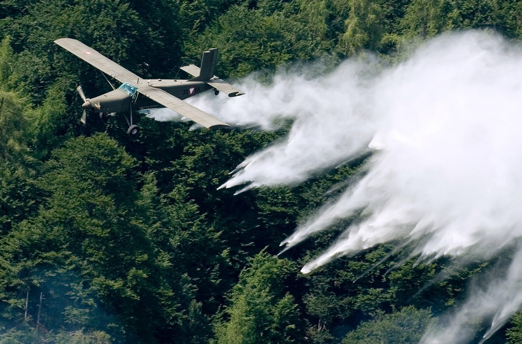 Ein olivfarbenes Militärflugzeug mit Propeller fliegt über einem dichten Wald und erzeugt mehrere weiße Rauchstreifen.