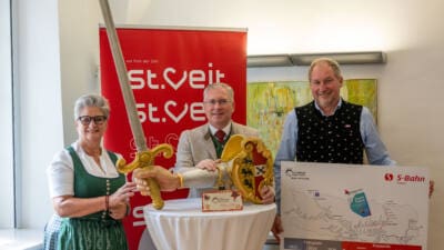 Eine Frau in Tracht und zwei Männer halten ein großes Schwert und eine Karte des Wiesenmarkts.