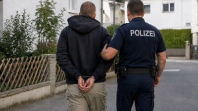 Polizeibeamter in Uniform führt gefesselten Mann mit Handschellen durch Wohnviertel
