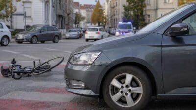Ein umgestürztes Fahrrad liegt neben einem grauen Pkw auf einer Straße in Graz, Polizeifahrzeug mit Blaulicht im Hintergrund