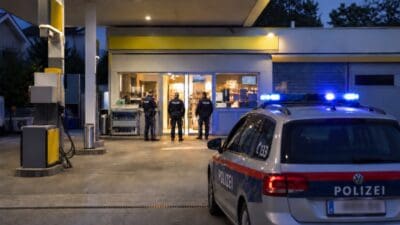 Polizeibeamte stehen vor einer beleuchteten Tankstelle mit einem Streifenwagen mit Blaulicht im Vordergrund bei Nacht