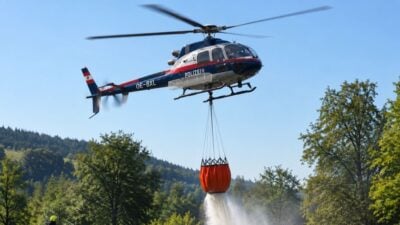 Ein Polizeihubschrauber fliegt über einer grünen Landschaft und lässt Wasser aus einem hängenden orangen Behälter herabfallen.