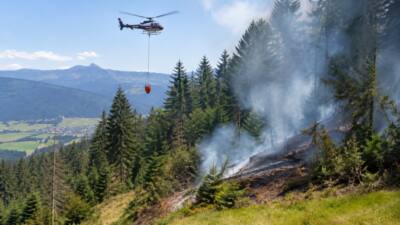 Ein Polizeihubschrauber mit hängendem Löschbehälter fliegt über einem verrauchten Bergwald.