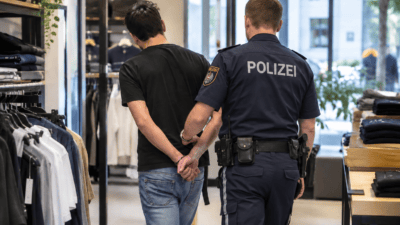 Polizist in Uniform führt einen Mann mit hinter dem Rücken gefesselten Händen durch ein Bekleidungsgeschäft.