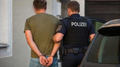 Polizist in Uniform führt jungen Mann mit Handschellen neben einem Auto ab.