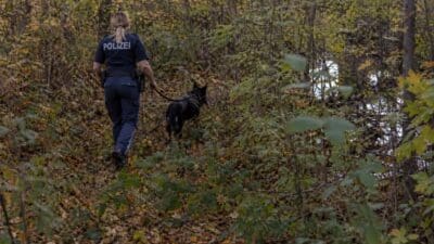 Polizistin in Uniform mit schwarzem Hund an Leine läuft bewaldeten Hang hinunter