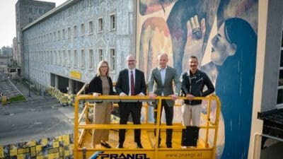 Vier Personen stehen auf einer gelben Hebebühne vor einer großen bunten Wandmalerei.