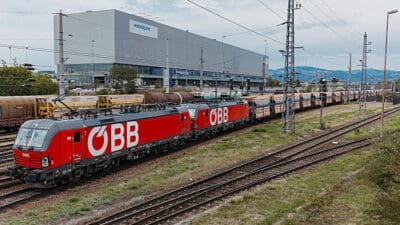 Rote ÖBB-Elektrolokomotive zieht Güterzug vor voestalpine-Gebäude auf Bahngleisen.
