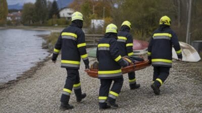 Vier Feuerwehrleute in dunkler Schutzkleidung mit gelben Streifen tragen eine verletzte Person auf einer Trage am Seeufer