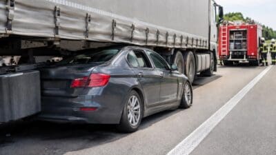 Ein grauer Pkw steckt unter dem Anhänger eines Sattelzugs auf der Autobahn, im Hintergrund ein rotes Feuerwehrfahrzeug.