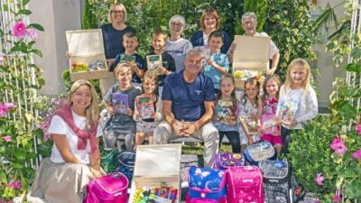 Erwachsene und Kinder posieren mit bunten Schulrucksäcken, Büchern und offenen Holzkisten voller Stifte im Freien.