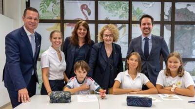 Fünf Erwachsene stehen hinter drei Kindern an einem Tisch in einem Klassenzimmer.