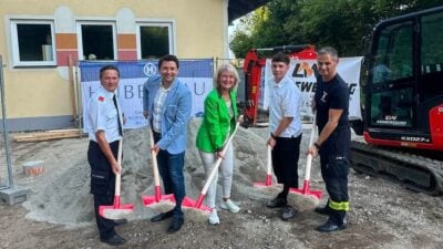 Fünf Personen halten Schaufeln mit Sand bei einer Spatenstichfeier auf einer Baustelle vor einem Gebäude.