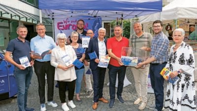 Zehn Erwachsene posieren vor blauem EU-Zelt und halten Fahnen sowie Informationshefte