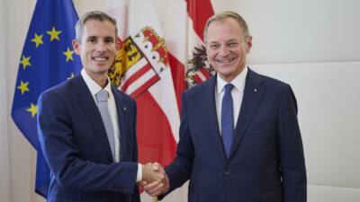 Stefan Blecha und Landeshauptmann Stelzer lächeln und schütteln sich die Hände vor Flaggen.