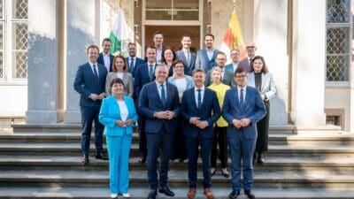 Gruppenporträt von zwanzig Frauen und Männern in Anzügen auf Steinstufen vor einem Gebäude mit Flaggen.