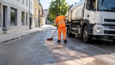 Ein Arbeiter in orangefarbener Warnkleidung kehrt eine nasse Straße neben einem weißen Tankwagen.
