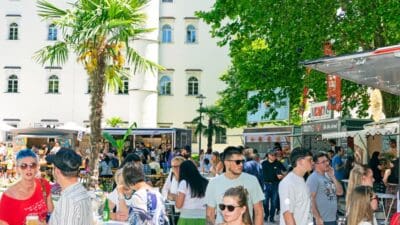 Eine Menschenmenge steht an einem sonnigen Street-Food-Markt mit Essensständen und Palmen vor einem weißen Gebäude