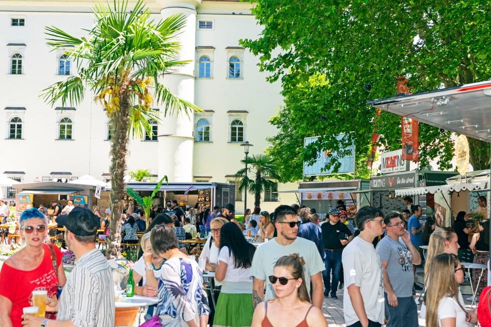 Eine Menschenmenge steht an einem sonnigen Street-Food-Markt mit Essensständen und Palmen vor einem weißen Gebäude