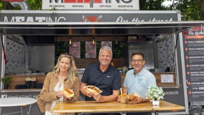Eine Frau und zwei Männer stehen vor einem Foodtruck und präsentieren Hotdogs sowie Hähnchenflügel.