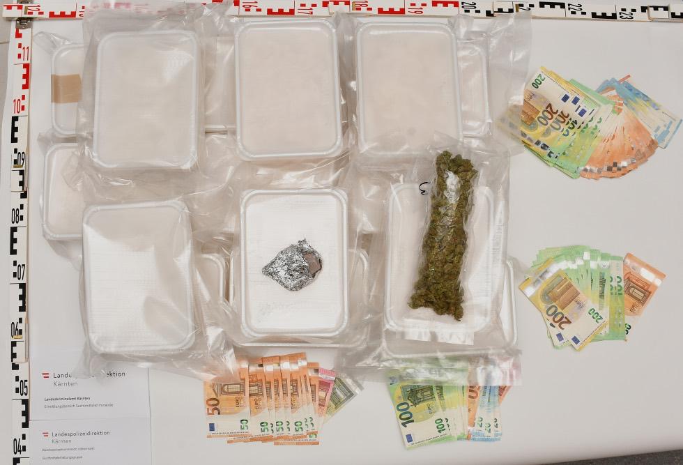 Sichergestellte Drogen in Plastikboxen, Cannabis und Bargeld auf einem Tisch mit Messskala
