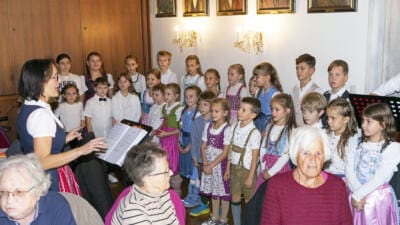 Eine Frau mit Notenheft dirigiert eine Gruppe von Kindern in Tracht bei einer Aufführung.