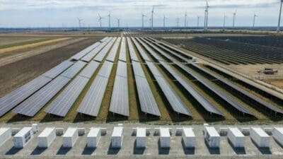 Ausgedehnte Photovoltaikanlage mit Reihen von Solarmodulen, weißen Batteriespeichern im Vordergrund und Windrädern im Hintergrund.