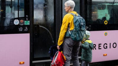 Ältere Frau in gelber Jacke mit Rucksack und Kind vor pinkem Bus