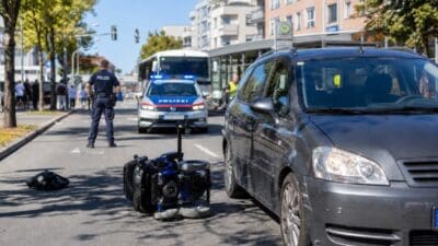 Umgestürzter Elektrorollstuhl liegt neben einem beschädigten Auto und einem Polizeifahrzeug mit Blaulicht auf einer Straße.