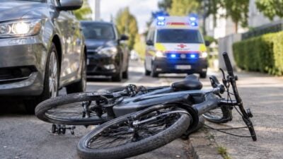 Umgestürztes Fahrrad liegt auf dem Asphalt vor parkenden Autos und einem Rettungswagen mit Blaulicht
