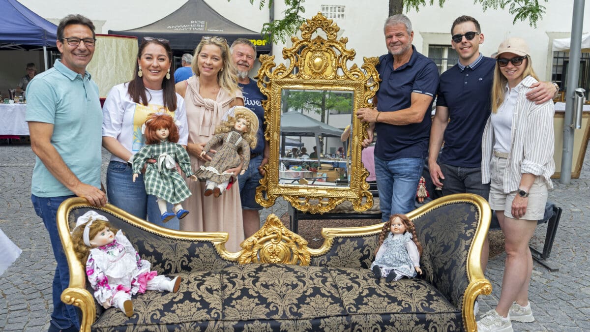 Sieben Personen stehen um ein antikes Sofa mit Puppen und einen großen goldenen Spiegel beim Flohmarkt.