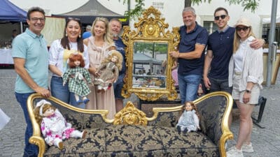 Sieben Personen stehen um ein antikes Sofa mit Puppen und einen großen goldenen Spiegel beim Flohmarkt.