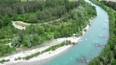 Luftaufnahme eines türkisfarbenen Flusses mit Kiesbänken und dicht bewaldetem Ufer bei Villach.